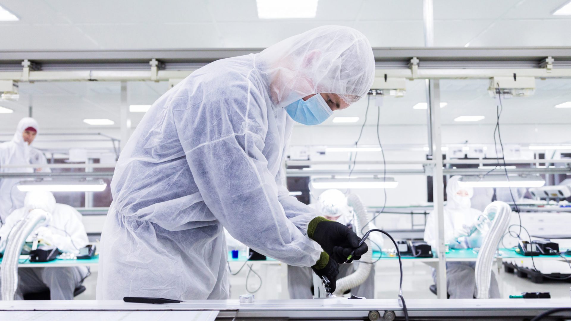 a factory worker in a white lab suit, black latex gloves and face mask, working with a soldering iron. other workers are on the background.